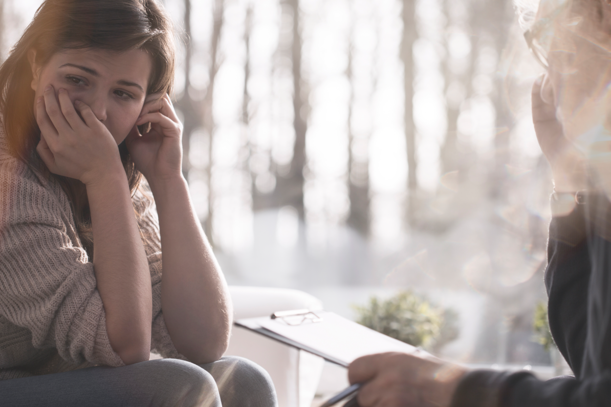 a person looks off to the side with her hand over her mouth and the other by her head; another person sits across from them with a notepad